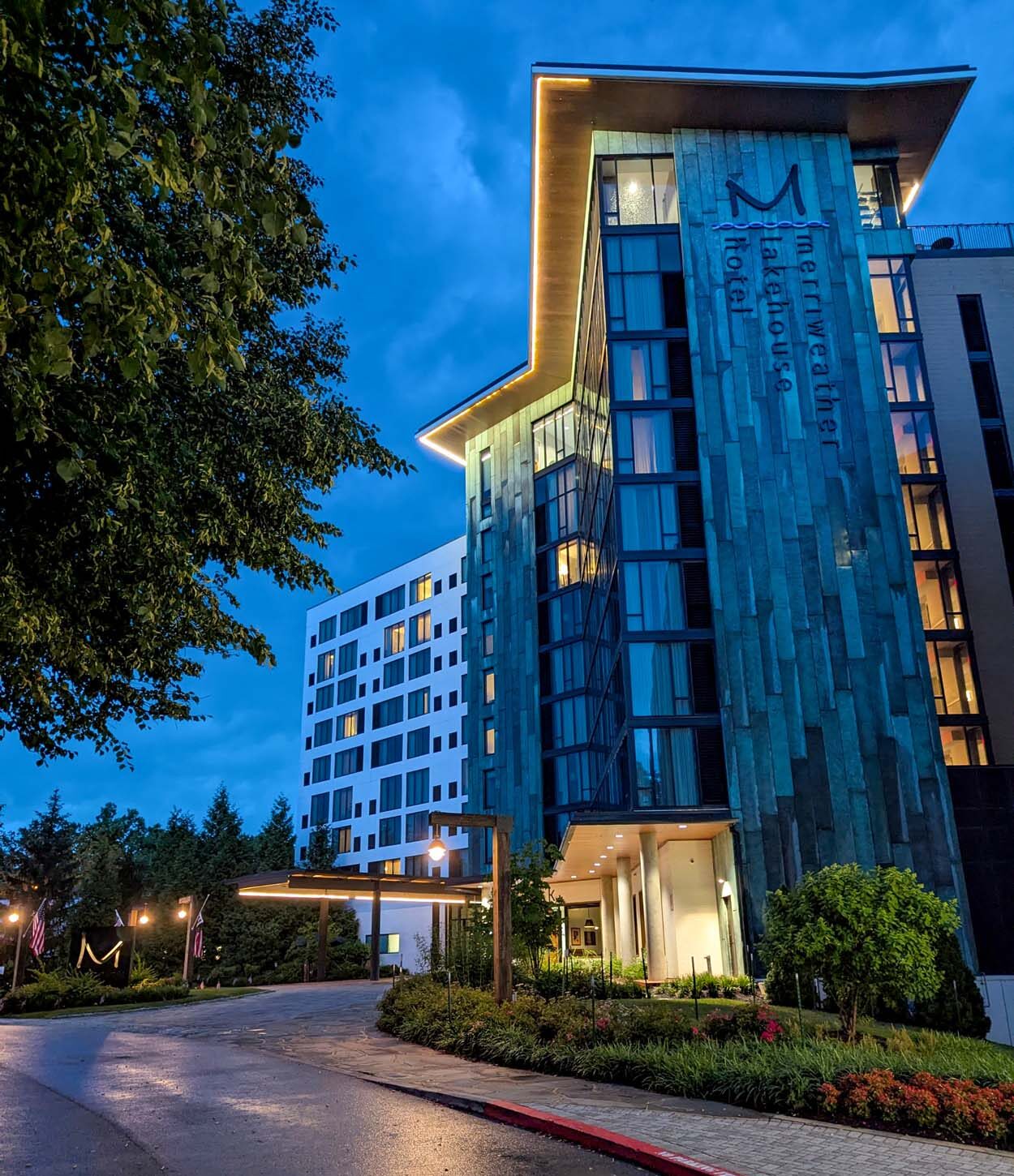 Merriweather Lakehouse Hotel at blue hour in Columbia Maryland with the illuminated Merriweather Lakehouse sign copper and glass facade glowing and the wet driveway reflecting the lights