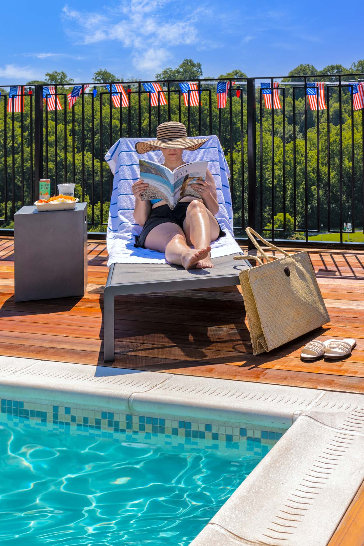 Guest in a wide brim straw hat reading on a lounge chair at the rooftop pool at Merriweather Lakehouse Hotel with American flag bunting strung along the railing and blue sky above