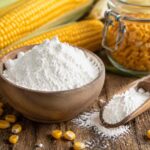 Wooden bowl filled with white cornstarch on a rustic wooden table, surrounded by fresh ears of corn, scattered corn kernels, a wooden scoop with cornstarch, and a glass jar of kernels in the background.
