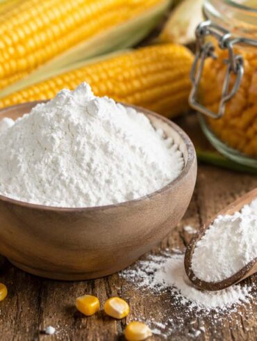 Wooden bowl filled with white cornstarch on a rustic wooden table, surrounded by fresh ears of corn, scattered corn kernels, a wooden scoop with cornstarch, and a glass jar of kernels in the background.