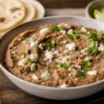A rustic ceramic bowl filled with creamy refried beans, topped with crumbled white cheese and fresh cilantro, sits on a wooden board beside warm flour tortillas, lime wedges, and diced onion in soft natural light.