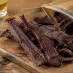 strips of dark, well-dried beef jerky arranged on a rustic wood cutting board, with a partially opened resealable kraft-paper bag beside it