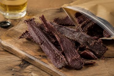 strips of dark, well-dried beef jerky arranged on a rustic wood cutting board, with a partially opened resealable kraft-paper bag beside it