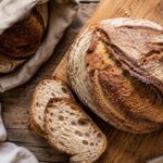 Overhead view of a rustic sourdough boule on a wooden cutting board with several sliced pieces revealing an open, airy crumb, beside a linen bread bag on a weathered wooden table in soft natural window light.