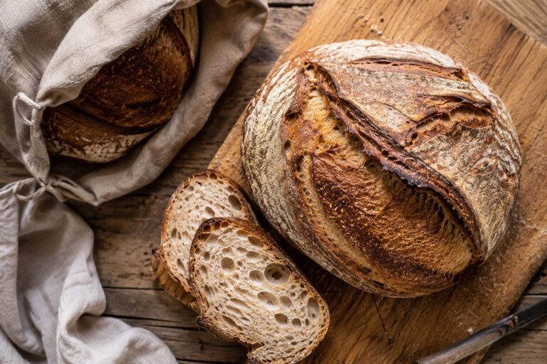 Overhead view of a rustic sourdough boule on a wooden cutting board with several sliced pieces revealing an open, airy crumb, beside a linen bread bag on a weathered wooden table in soft natural window light.