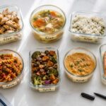 neatly organized glass food storage containers holding various leftovers including chicken, soup, rice, and pasta, arranged on a clean white surface with handwritten date labels.