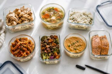 neatly organized glass food storage containers holding various leftovers including chicken, soup, rice, and pasta, arranged on a clean white surface with handwritten date labels.
