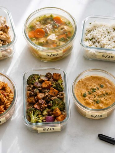 neatly organized glass food storage containers holding various leftovers including chicken, soup, rice, and pasta, arranged on a clean white surface with handwritten date labels.