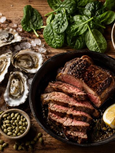 A rustic wooden table scattered with a curated spread of iron-rich foods: a small cast iron skillet with seared lean beef and a lemon wedge, a handful of raw oysters on the half shell with crushed ice, a bowl of cooked lentils topped with fresh cherry tomatoes, a small pile of fresh spinach leaves, a few squares of dark chocolate, a scoop of pumpkin seeds, and a wedge of lemon cut in half