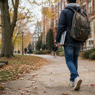 Young college student walking across a tree-lined university campus in autumn, carrying books and a backpack along a leaf-covered pathway with historic brick academic buildings in the background.