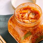 A jar of traditional homemade kimchi on a marble surface with wooden chopsticks beside it.