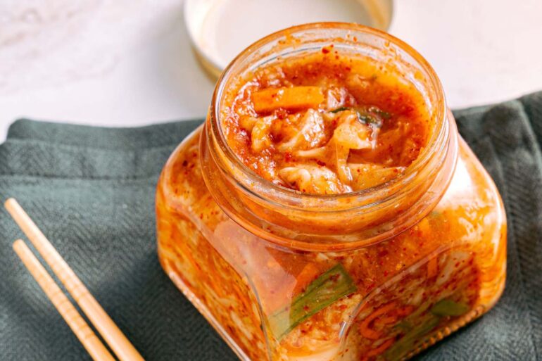 A jar of traditional homemade kimchi on a marble surface with wooden chopsticks beside it.
