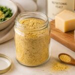 An open glass jar filled with golden nutritional yeast flakes sits on a bright kitchen countertop beside a small wooden spoon and scattered flakes. Softly blurred ingredients in the background, including a salad, cashews, and a wedge of cheese, create a realistic editorial food scene illustrating nutritional yeast and proper pantry storage.
