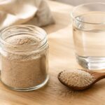 A small glass jar filled with pale beige psyllium husk sits open on a light wooden countertop, with a wooden spoon holding a small mound of psyllium husk beside it and a clear glass of water nearby.
