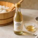 A bottle of rice vinegar sits beside a traditional wooden sushi rice tub filled with freshly cooked rice, with a small bowl of rice vinegar and a wooden rice paddle nearby. The bright, minimalist food scene highlights rice vinegar as a key ingredient for seasoning sushi rice.