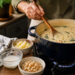 A home cook stirs a pot of soup on the stove while preparing it without cream. Nearby are the substitute ingredients in small bowls: a cornstarch slurry, diced potato, canned white beans, rice, rolled oats, silken tofu, canned coconut milk, and egg yolks, illustrating practical ways to thicken soup without cream in a bright, natural kitchen.