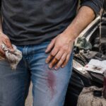 A man works on a car in a driveway with reddish transmission fluid staining the thigh of his blue jeans, while his oily hands, a used rag, and an unbranded bottle of transmission fluid are visible beside the open hood.