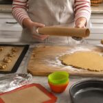 A home baker holds an empty parchment paper roll over a kitchen counter while preparing cookie dough and pie crust. Nearby are a silicone baking mat with cookie dough balls, aluminum foil, reusable silicone muffin liners, a greased cake pan, and a rolling pin, illustrating practical parchment paper substitutes for baking and food prep.