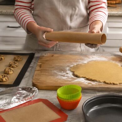 A home baker holds an empty parchment paper roll over a kitchen counter while preparing cookie dough and pie crust. Nearby are a silicone baking mat with cookie dough balls, aluminum foil, reusable silicone muffin liners, a greased cake pan, and a rolling pin, illustrating practical parchment paper substitutes for baking and food prep.