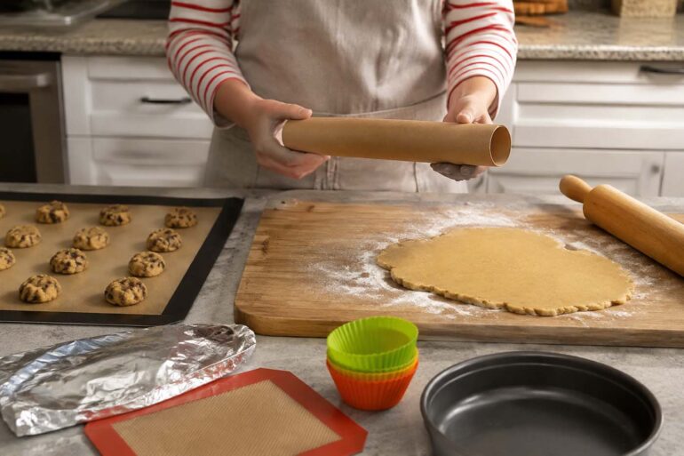 A home baker holds an empty parchment paper roll over a kitchen counter while preparing cookie dough and pie crust. Nearby are a silicone baking mat with cookie dough balls, aluminum foil, reusable silicone muffin liners, a greased cake pan, and a rolling pin, illustrating practical parchment paper substitutes for baking and food prep.