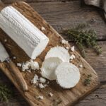A log of fresh goat cheese with several sliced rounds, crumbs, and thyme arranged on a rustic wooden cutting board beside a small cheese knife, photographed from above on a weathered wood table.