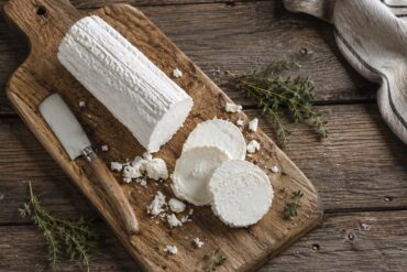 A log of fresh goat cheese with several sliced rounds, crumbs, and thyme arranged on a rustic wooden cutting board beside a small cheese knife, photographed from above on a weathered wood table.