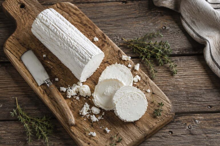 A log of fresh goat cheese with several sliced rounds, crumbs, and thyme arranged on a rustic wooden cutting board beside a small cheese knife, photographed from above on a weathered wood table.