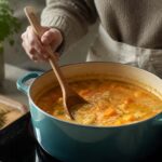 Home cook stirring a pot of chunky squash soup with a wooden spoon in a teal Dutch oven, with fresh herbs and vegetables nearby.