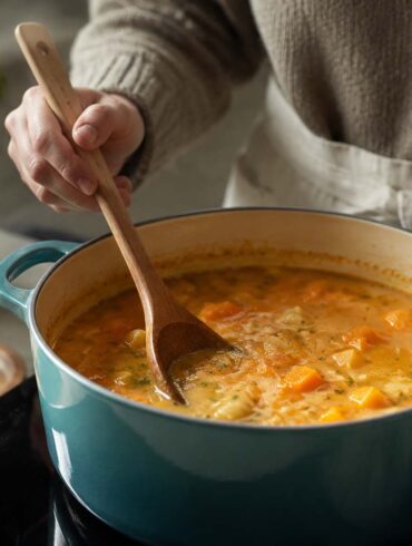 Home cook stirring a pot of chunky squash soup with a wooden spoon in a teal Dutch oven, with fresh herbs and vegetables nearby.