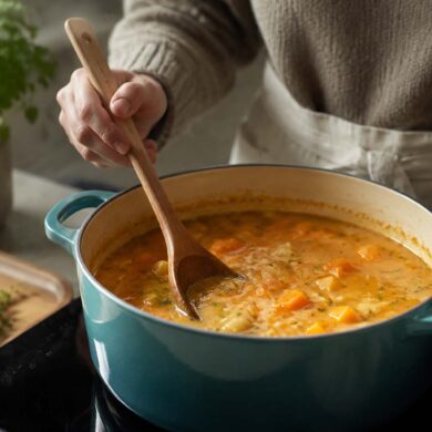 Home cook stirring a pot of chunky squash soup with a wooden spoon in a teal Dutch oven, with fresh herbs and vegetables nearby.