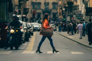 A busy urban street in Rome, Italy, with a young woman crossing among cars and pedestrians.
