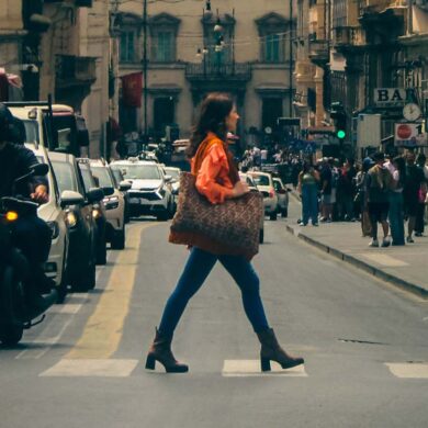A busy urban street in Rome, Italy, with a young woman crossing among cars and pedestrians.