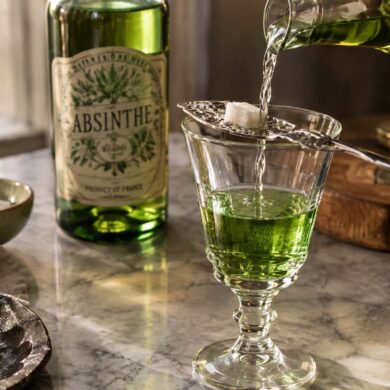 A moody still life shows a vivid green absinthe-style drink being prepared in a clear stemmed glass on a marble tabletop. A stream of clear liquid is being poured over a white sugar cube resting on an ornate slotted absinthe spoon across the glass, while a green glass bottle labeled “ABSINTHE” stands softly out of focus in the background. A small bowl of sugar cubes, green olives on a dark plate, folded linen, old books, a wooden serving board, a small vase of flowers, and a candle surround the scene, with warm natural light creating an intimate vintage atmosphere.