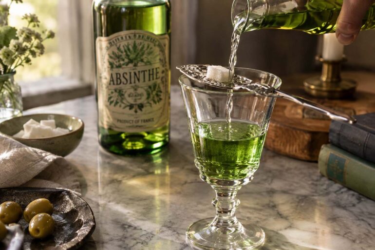 A moody still life shows a vivid green absinthe-style drink being prepared in a clear stemmed glass on a marble tabletop. A stream of clear liquid is being poured over a white sugar cube resting on an ornate slotted absinthe spoon across the glass, while a green glass bottle labeled “ABSINTHE” stands softly out of focus in the background. A small bowl of sugar cubes, green olives on a dark plate, folded linen, old books, a wooden serving board, a small vase of flowers, and a candle surround the scene, with warm natural light creating an intimate vintage atmosphere.