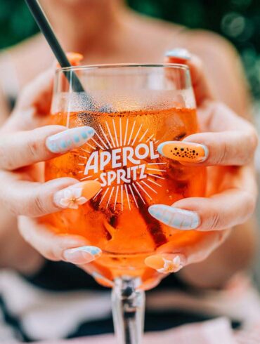 Close-up of a woman enjoying an Aperol Spritz cocktail outdoors on a sunny day, showcasing vibrant nail art.