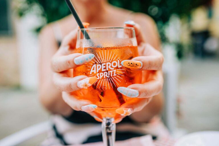 Close-up of a woman enjoying an Aperol Spritz cocktail outdoors on a sunny day, showcasing vibrant nail art.