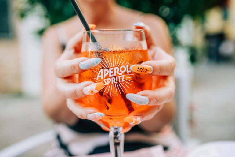Close-up of a woman enjoying an Aperol Spritz cocktail outdoors on a sunny day, showcasing vibrant nail art.