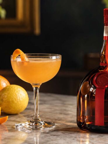 A citrus-colored cocktail in a coupe glass sits beside a dark amber liqueur bottle with a red ribbon and cap, surrounded by oranges, a lemon, and an orange peel on a marble counter. Warm natural light creates an elegant evening bar setting.