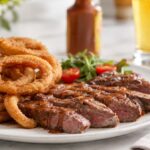 Close-up of sliced medium-rare steak drizzled with brown sauce and served with golden fried onion rings on a white plate, with a red-capped bottle of brown steak sauce softly blurred in the background on a bright kitchen table.