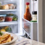 Open refrigerator stocked with fresh produce, eggs, cheese, and leftovers, with a red-capped bottle of brown steak sauce standing in the refrigerator door shelf and a plate of steak and fries softly blurred in the foreground.