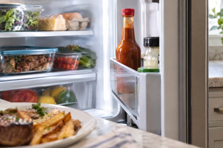 Open refrigerator stocked with fresh produce, eggs, cheese, and leftovers, with a red-capped bottle of brown steak sauce standing in the refrigerator door shelf and a plate of steak and fries softly blurred in the foreground.