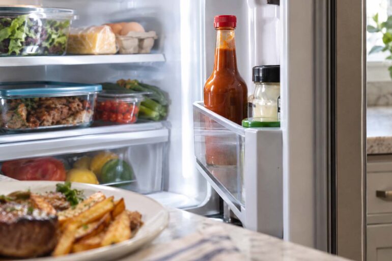 Open refrigerator stocked with fresh produce, eggs, cheese, and leftovers, with a red-capped bottle of brown steak sauce standing in the refrigerator door shelf and a plate of steak and fries softly blurred in the foreground.