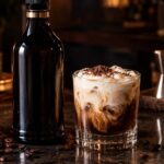 a KahlĂşa-style coffee liqueur bottle beside a White Russian cocktail with cream swirled on top, dark moody bar lighting, coffee beans scattered nearby,
