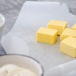 Close-up of margarine cubes on parchment paper with bowls of ingredients in a kitchen baking setting