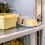 A tub of margarine on a refrigerator shelf next to a stick of margarine in its butter dish, refrigerator door open in the background