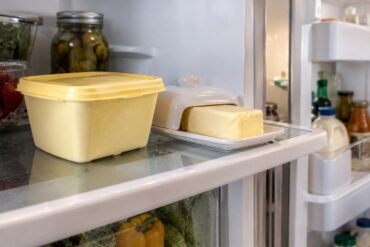 A tub of margarine on a refrigerator shelf next to a stick of margarine in its butter dish, refrigerator door open in the background