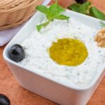 A mouth-watering tzatziki dip garnished with olives, mint, and walnuts on a wooden table