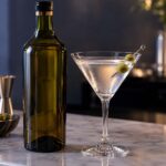 a vermouth bottle beside a classic martini glass with an olive garnish on a marble bar counter, soft cool evening lighting