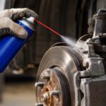 Close-up view inside a vehicle’s wheel well showing a gloved hand holding a plain blue aerosol spray can with a thin red straw nozzle. A fine mist is being sprayed from the straw onto a dusty, weathered brake caliper and brake rotor. The exposed wheel hub and several wheel studs are visible in the foreground, with rust and grime on the surrounding metal components. The background is softly blurred, emphasizing the can, spray, and brake assembly.