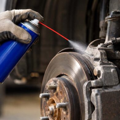 Close-up view inside a vehicle’s wheel well showing a gloved hand holding a plain blue aerosol spray can with a thin red straw nozzle. A fine mist is being sprayed from the straw onto a dusty, weathered brake caliper and brake rotor. The exposed wheel hub and several wheel studs are visible in the foreground, with rust and grime on the surrounding metal components. The background is softly blurred, emphasizing the can, spray, and brake assembly.